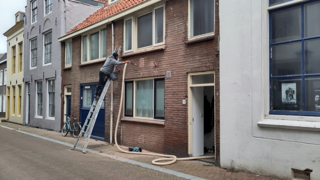 Foto van een gevel, ladder ertegen met een man erop die een beige slang in een gaatje in de muur steekt Foto van een gevel, ladder ertegen met een man erop die een beige slang in een gaatje in de muur steekt