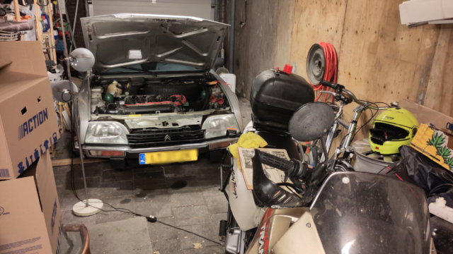 Grijze Citroën CX in de garage. Motorkap open, een schemerlamp ernaast die de motorruimte verlicht. Ervoor staat een motorfiets met een motorhelm ernaast. Grijze Citroën CX in de garage. Motorkap open, een schemerlamp ernaast die de motorruimte verlicht. Ervoor staat een motorfiets met een motorhelm ernaast.