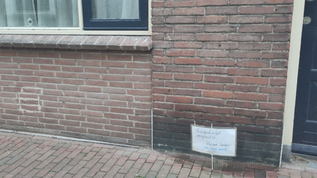 Foto van een stuk voorgevel, groen gaas ervoor en een bordje erop met 'hondentoilet verplaatst, nieuwe lokatie: uw eigen gevel - lach naar het vogeltje: camerabewaking'. Linksboven is een webcam achter het raam te zien. Foto van een stuk voorgevel, groen gaas ervoor en een bordje erop met 'hondentoilet verplaatst, nieuwe lokatie: uw eigen gevel - lach naar het vogeltje: camerabewaking'. Linksboven is een webcam achter het raam te zien.