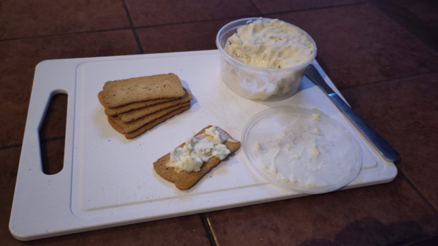Witte snijplank op tafel, met daarop een stapeltje melba-toastjes, een met gorgonzola. Ernaast een rond plastic bakje vol gorgonzola. Witte snijplank op tafel, met daarop een stapeltje melba-toastjes, een met gorgonzola. Ernaast een rond plastic bakje vol gorgonzola.
