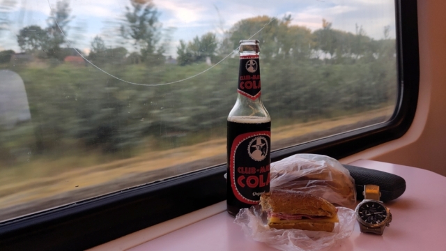  Foto vanuit de trein: op de voorgrond twee broodjes, een flesje Club-Mate cola, een horloge en brillenkoker. Door het raam zien we het Zeeuwse landschap, onscherp omdat de trein rijdt.